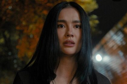 Young woman with long black hair in a dark top stands outdoors at night, illuminated by blurred city lights in the background.