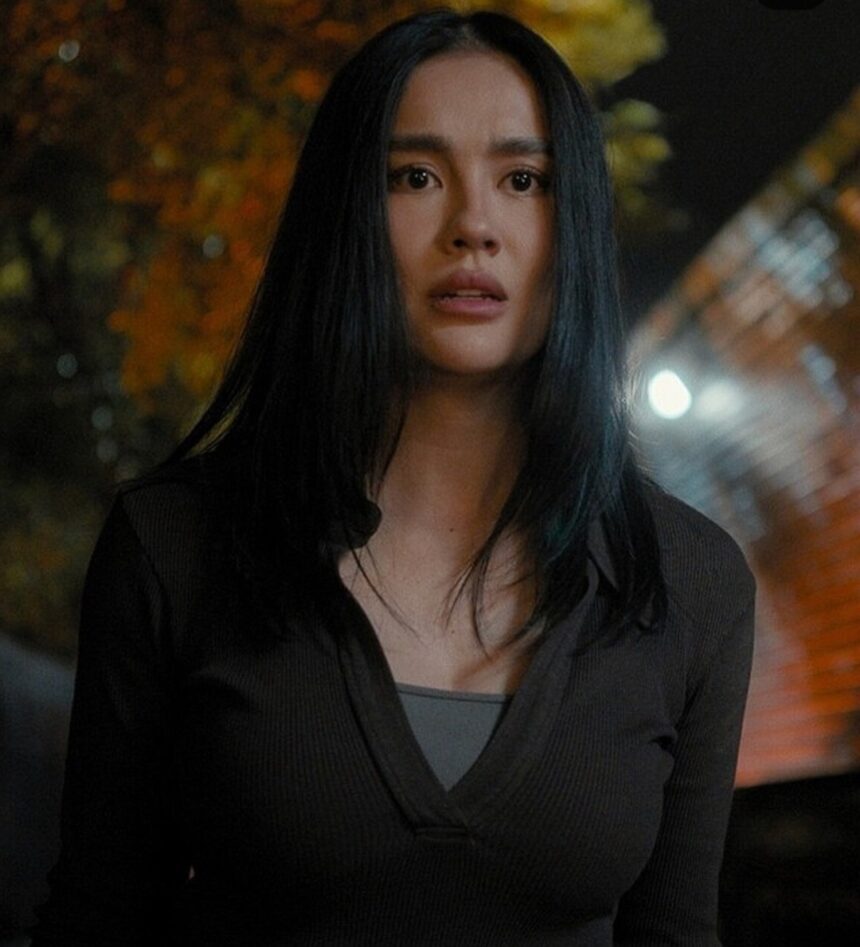 Young woman with long black hair in a dark top stands outdoors at night, illuminated by blurred city lights in the background.