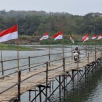 A bamboo pedestrian bridge over a calm river with red-and-white flags along the railing; a person on a scooter crosses the bridge, trees in the background.