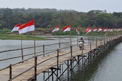 A bamboo pedestrian bridge over a calm river with red-and-white flags along the railing; a person on a scooter crosses the bridge, trees in the background.