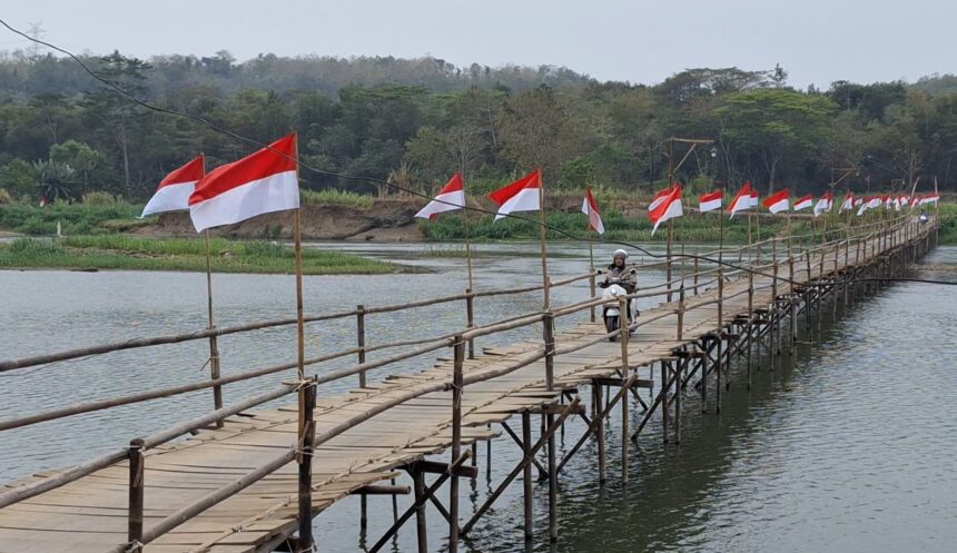A bamboo pedestrian bridge over a calm river with red-and-white flags along the railing; a person on a scooter crosses the bridge, trees in the background.