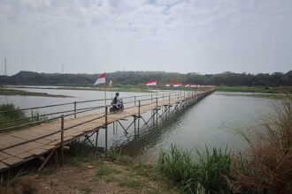 Rider on a scooter crosses a long bamboo bridge over a calm river, with Indonesian flags lining the railing and a rural landscape in the background.