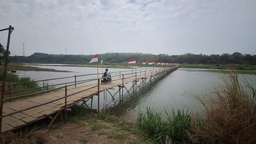 Rider on a scooter crosses a long bamboo bridge over a calm river, with Indonesian flags lining the railing and a rural landscape in the background.