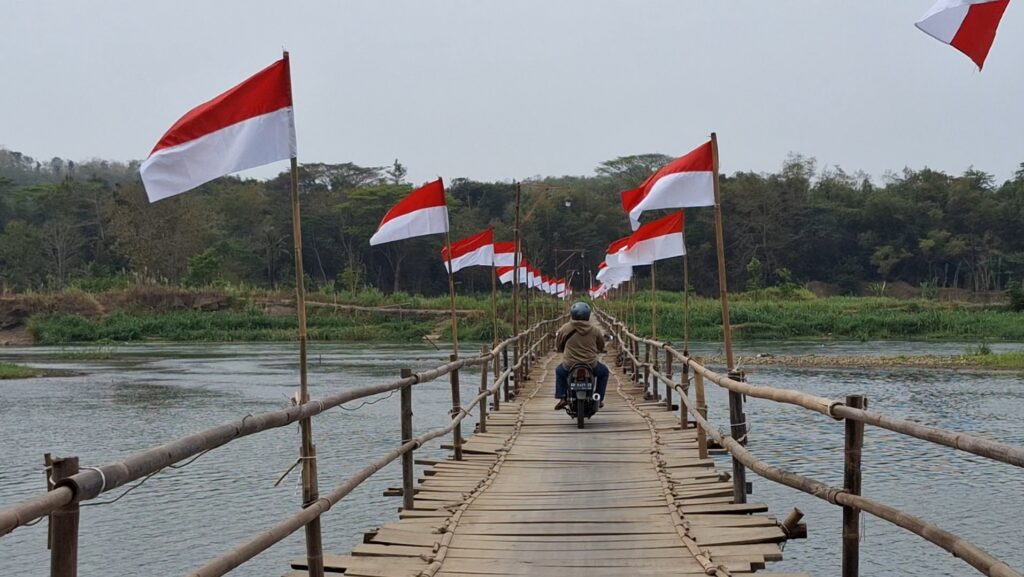 Screenshot_20260811_090614_Gallery - Mabur.co Motorcyclist crossing a long bamboo bridge lined with red-and-white flags over a calm river: rural scene with trees in the background.