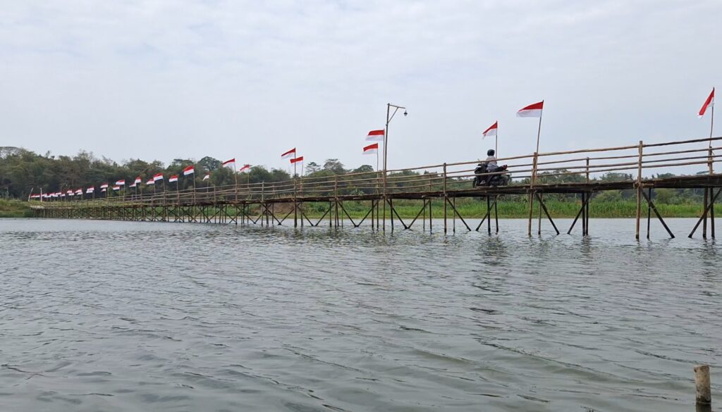 Long wooden bridge over a river with red-and-white flags; a motorcyclist rides across.