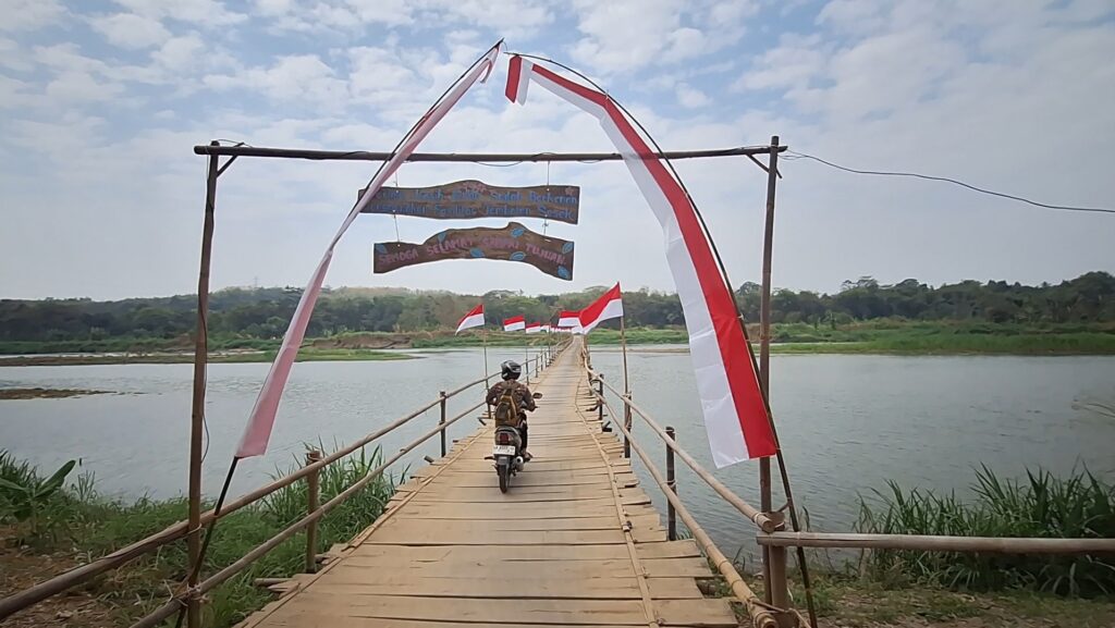 Screenshot_20260811_090647_Gallery - Mabur.co Bamboo pedestrian bridge over a calm river with red-and-white flags along the sides; a motorbike rides toward banners overhead.