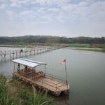 Wooden pedestrian bridge extending over a calm river toward a tree-lined shore, flags along the path and a small raft with a covered seating area nearby (Indonesia flag).