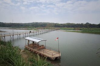 Wooden pedestrian bridge extending over a calm river toward a tree-lined shore, flags along the path and a small raft with a covered seating area nearby (Indonesia flag).