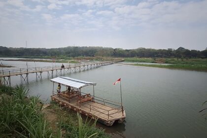 Wooden pedestrian bridge extending over a calm river toward a tree-lined shore, flags along the path and a small raft with a covered seating area nearby (Indonesia flag).