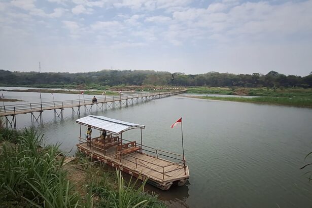 Wooden pedestrian bridge extending over a calm river toward a tree-lined shore, flags along the path and a small raft with a covered seating area nearby (Indonesia flag).
