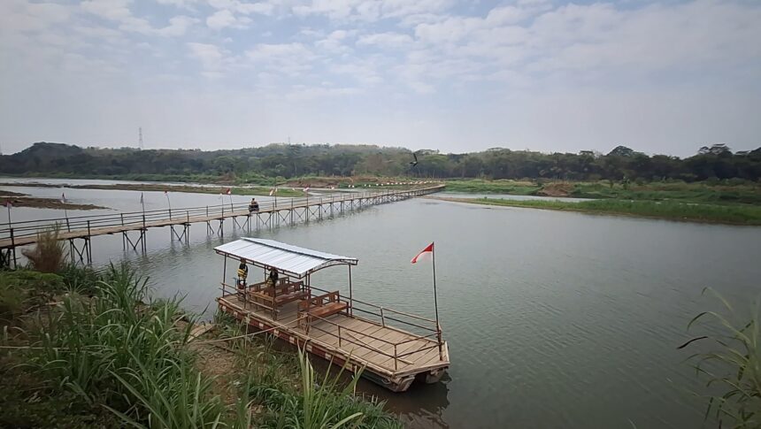 Wooden pedestrian bridge extending over a calm river toward a tree-lined shore, flags along the path and a small raft with a covered seating area nearby (Indonesia flag).