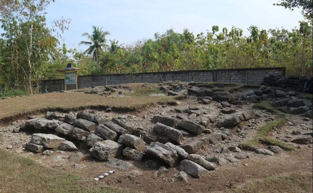A rocky excavation area with scattered limestone blocks and cleared earth, a low wall in the background and trees beyond.