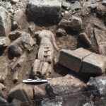 Archaeological dig scene with rough stones and dirt; a partially exposed carved stone resembling a face amid the rubble, with measurement scales nearby and a marker plate in the corner.