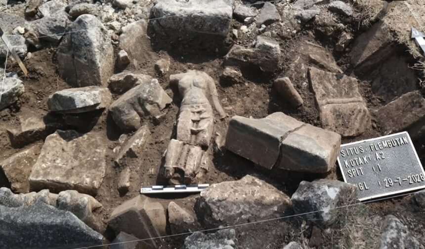 Archaeological dig scene with rough stones and dirt; a partially exposed carved stone resembling a face amid the rubble, with measurement scales nearby and a marker plate in the corner.