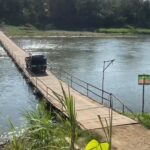 A dark SUV driving on a narrow wooden bridge across a calm river, with green vegetation along the far bank.
