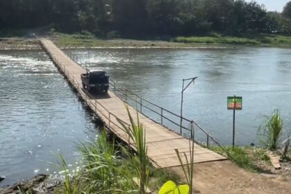 A dark SUV driving on a narrow wooden bridge across a calm river, with green vegetation along the far bank.
