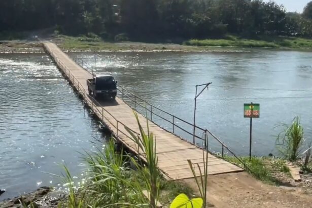 A dark SUV driving on a narrow wooden bridge across a calm river, with green vegetation along the far bank.