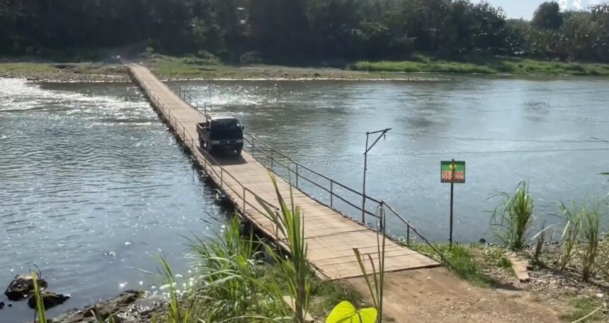 A dark SUV driving on a narrow wooden bridge across a calm river, with green vegetation along the far bank.