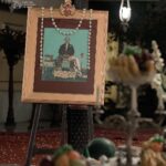 Framed ceremonial portrait on an easel with a flower garland, in a dimly lit ritual setting with offerings in the foreground.