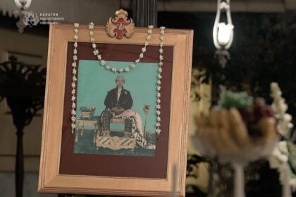 Framed ceremonial portrait on an easel with a flower garland, in a dimly lit ritual setting with offerings in the foreground.