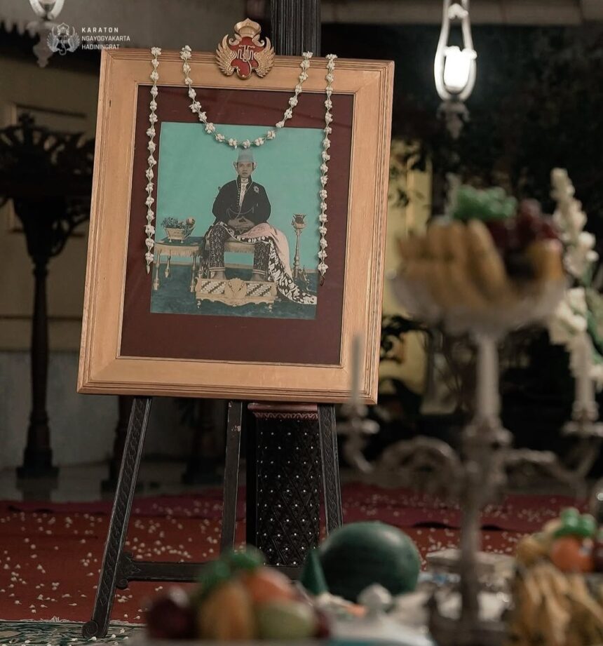 Framed ceremonial portrait on an easel with a flower garland, in a dimly lit ritual setting with offerings in the foreground.