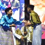 Two men in traditional Malay attire greet each other with a handshake on stage during a cultural performance, with a colorful backdrop behind them.