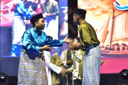 Two men in traditional Malay attire greet each other with a handshake on stage during a cultural performance, with a colorful backdrop behind them.