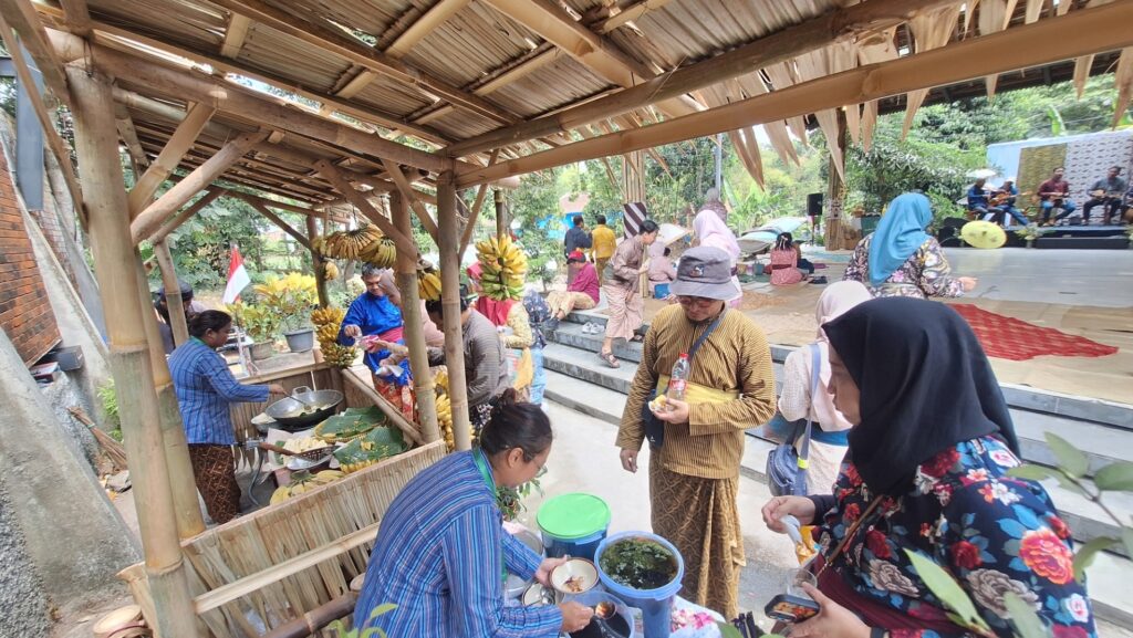 Outdoor cultural festival with bamboo food stalls, banana decorations, and people in traditional clothing queuing for snacks near the stall, stage with musicians in the background.