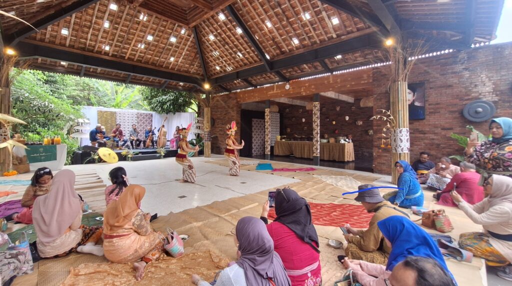 Outdoor-style hall with a wooden roof hosting a traditional dance performance; two dancers in red and gold costumes perform while a live band plays in the background, audience seated on woven mats around the stage.