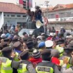 Large crowd of protesters surrounds a car on a street; people stand on the car's roof as a megaphone sits nearby, guarded by police.