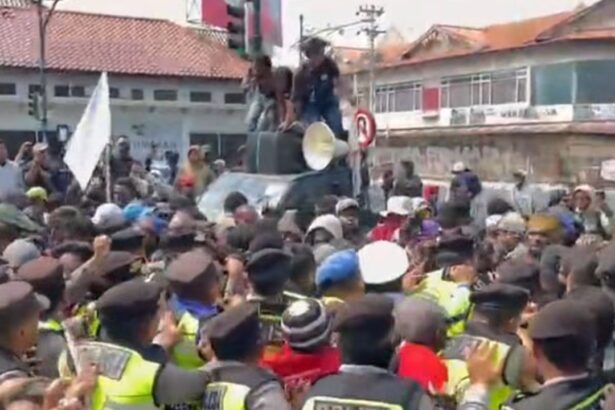 Large crowd of protesters surrounds a car on a street; people stand on the car's roof as a megaphone sits nearby, guarded by police.