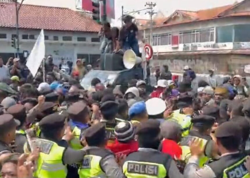 Large crowd of protesters surrounds a car on a street; people stand on the car's roof as a megaphone sits nearby, guarded by police.
