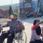 At an outdoor festival, a man in a wheelchair and two kids hold a green ribbon while one child bites a hanging pastry, with onlookers and a mural backdrop in the background.