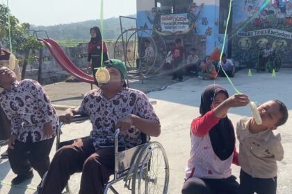At an outdoor festival, a man in a wheelchair and two kids hold a green ribbon while one child bites a hanging pastry, with onlookers and a mural backdrop in the background.