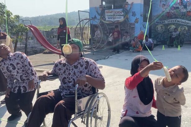 At an outdoor festival, a man in a wheelchair and two kids hold a green ribbon while one child bites a hanging pastry, with onlookers and a mural backdrop in the background.