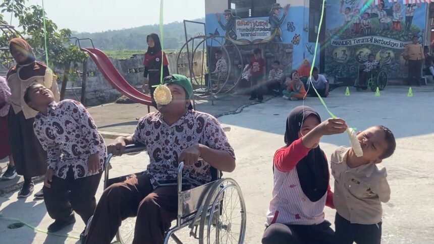 At an outdoor festival, a man in a wheelchair and two kids hold a green ribbon while one child bites a hanging pastry, with onlookers and a mural backdrop in the background.