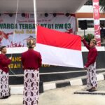 Four students in traditional Indonesian clothing raise a large red-and-white flag on a stage backdrop celebrating RI’s 81st anniversary.