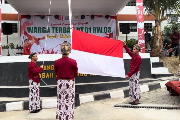 Four students in traditional Indonesian clothing raise a large red-and-white flag on a stage backdrop celebrating RI’s 81st anniversary.
