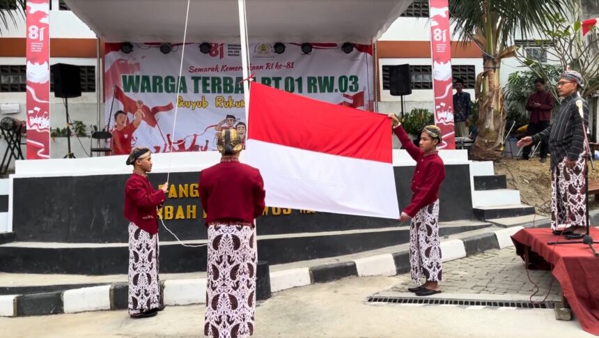 Four students in traditional Indonesian clothing raise a large red-and-white flag on a stage backdrop celebrating RI’s 81st anniversary.