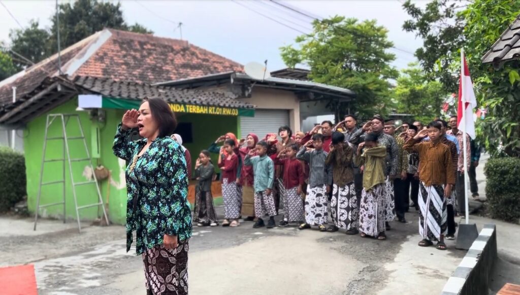 A woman in a blue floral dress leads a crowd in batik clothing saluting on a village street, with a green house in the background.