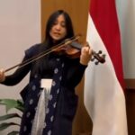 Violinist performing on stage, bow drawn across the violin, wearing a dark outfit with a snowflake-pattern scarf; a red‑and‑white flag behind her.