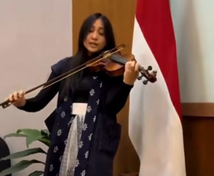 Violinist performing on stage, bow drawn across the violin, wearing a dark outfit with a snowflake-pattern scarf; a red‑and‑white flag behind her.