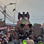 Large demon-like float with a dragon on its back in a street parade, crowds watching along the street.