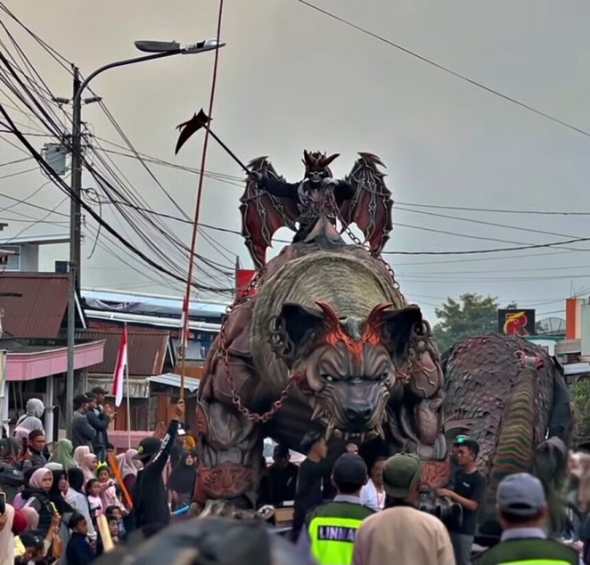 Large demon-like float with a dragon on its back in a street parade, crowds watching along the street.