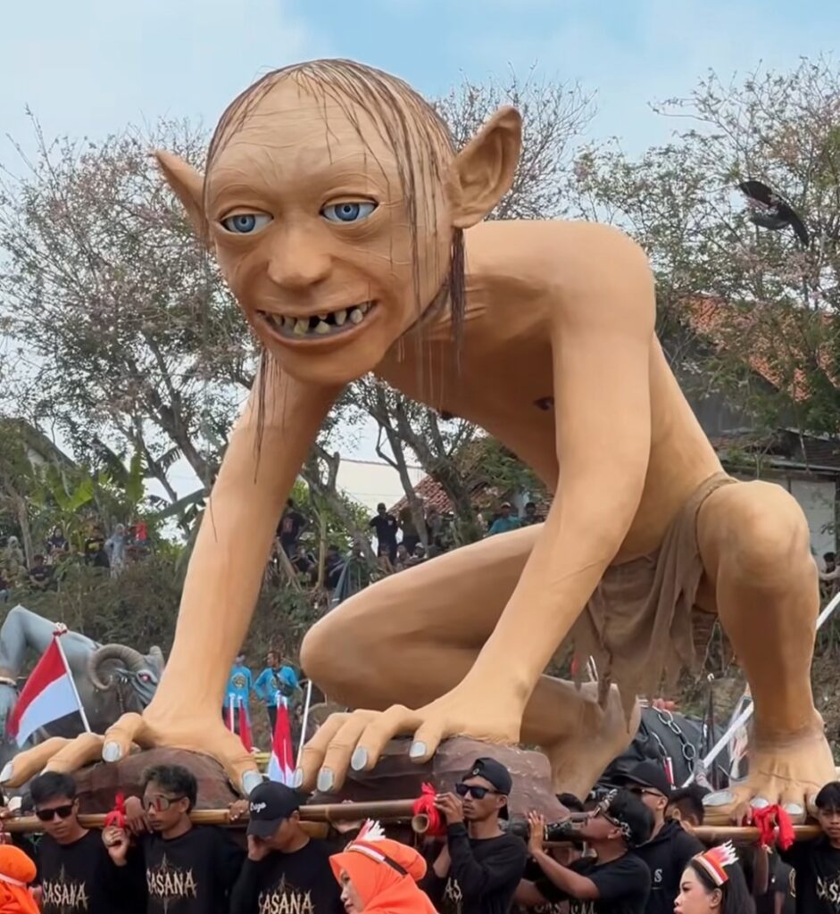 Screenshot_20260818_125839_Instagram - Mabur.co Giant humanoid puppet with pale skin and blue eyes crouches on a platform carried by performers at a festival.