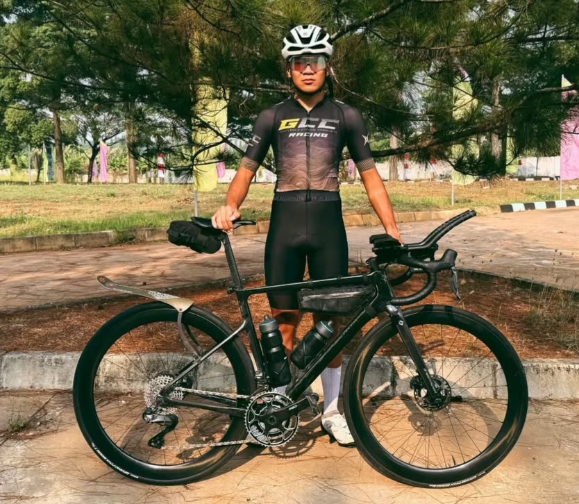 Cyclist in black racing kit and helmet standing beside a black road bike outdoors on a sunny day.