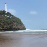 Lighthouse atop a rocky, tree-covered cliff overlooking a sandy beach and gentle ocean waves.