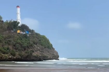 Lighthouse atop a rocky, tree-covered cliff overlooking a sandy beach and gentle ocean waves.