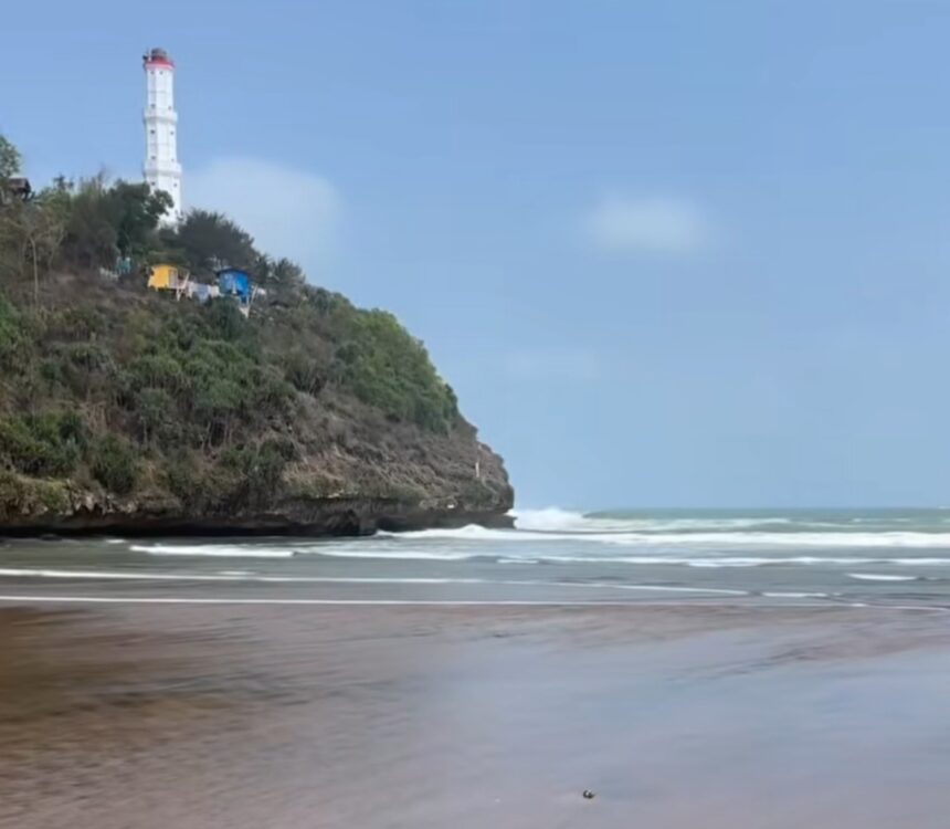 Lighthouse atop a rocky, tree-covered cliff overlooking a sandy beach and gentle ocean waves.