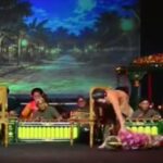 Onstage, a dancer lies on the floor beside a standing partner while a live band sits in the background.
